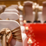 Close up of gold ribbons on white chair covers, blurred red carpet aisle in background
